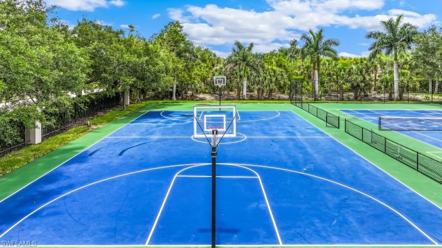 Full-size basketball court featuring a blue playing surface with white line markings, two basketball hoops, surrounding green perimeter, and mature landscaping