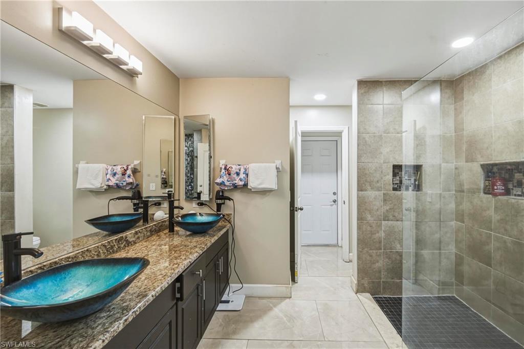 Spacious bathroom featuring a dual vanity with vessel sinks, a full-width mirror, a tiled walk-in shower with glass enclosure, recessed lighting, and tile flooring