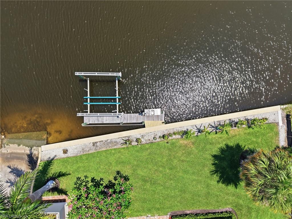 Waterfront property featuring a private boat lift, a boat dock, a concrete seawall, a manicured lawn, and mature tropical landscaping