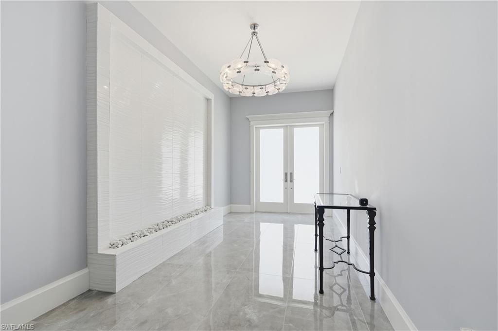 Hallway featuring a built-in water wall with textured white tile and decorative stones, double-door entry, overhead chandelier, and polished stone tile flooring