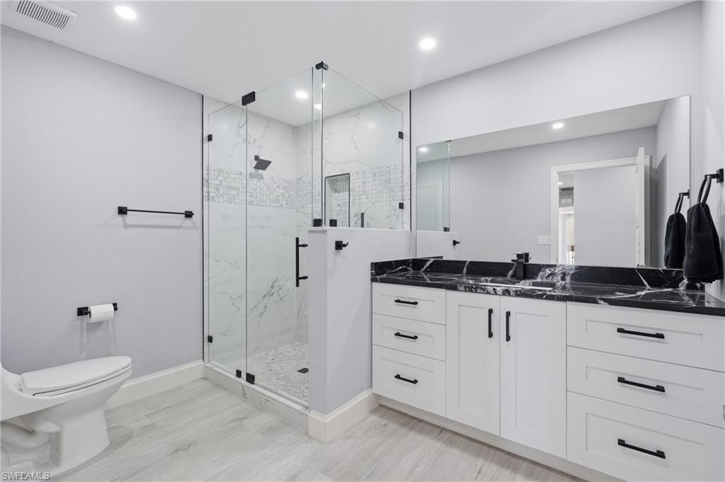 Contemporary bathroom featuring a spacious glass-enclosed shower with white marble-look tile, a dual vanity with a black stone countertop, white shaker-style cabinetry, and light wood-finish flooring