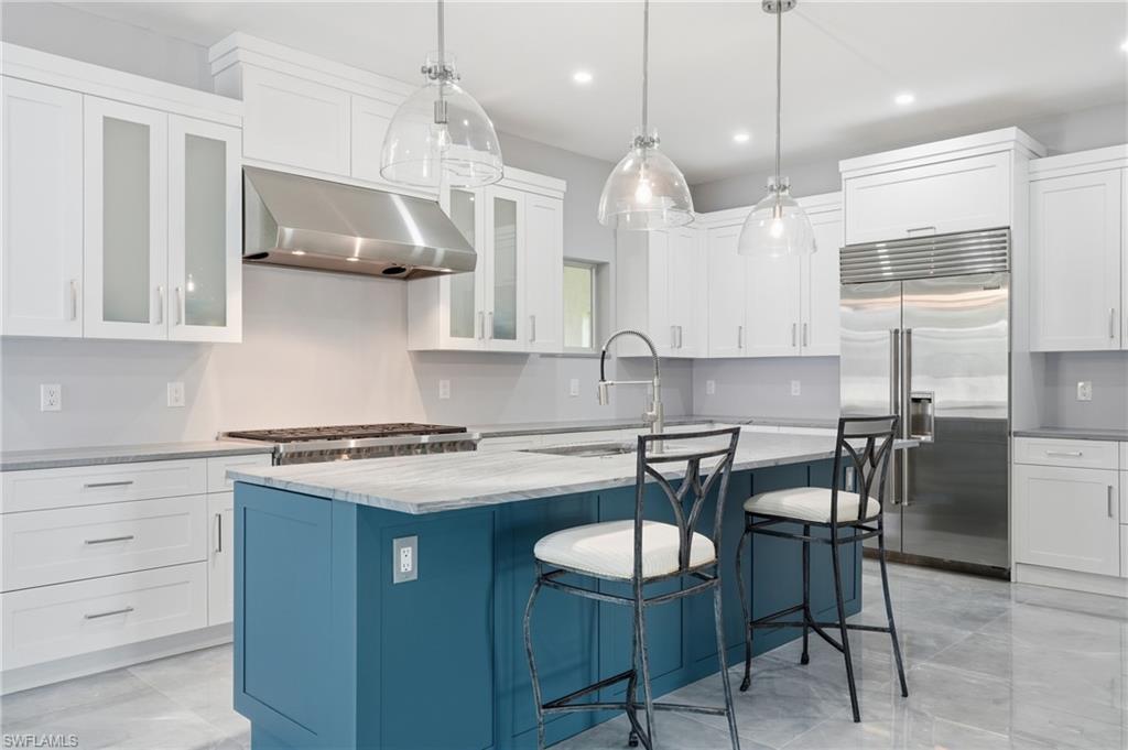 Kitchen featuring a blue island with a contrasting stone countertop, clear pendant lighting, stainless steel appliances, white cabinetry with some frosted glass doors, and light-toned tile flooring