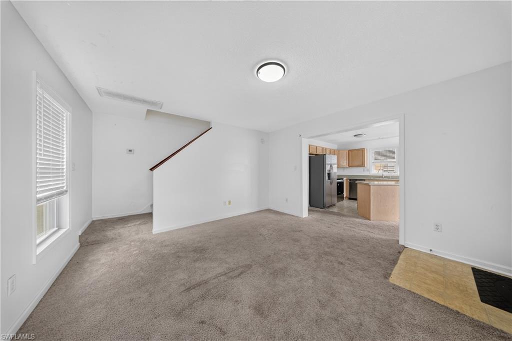 Open-concept living space featuring neutral carpeting, white walls, and a staircase with a dark wood-finish handrail
