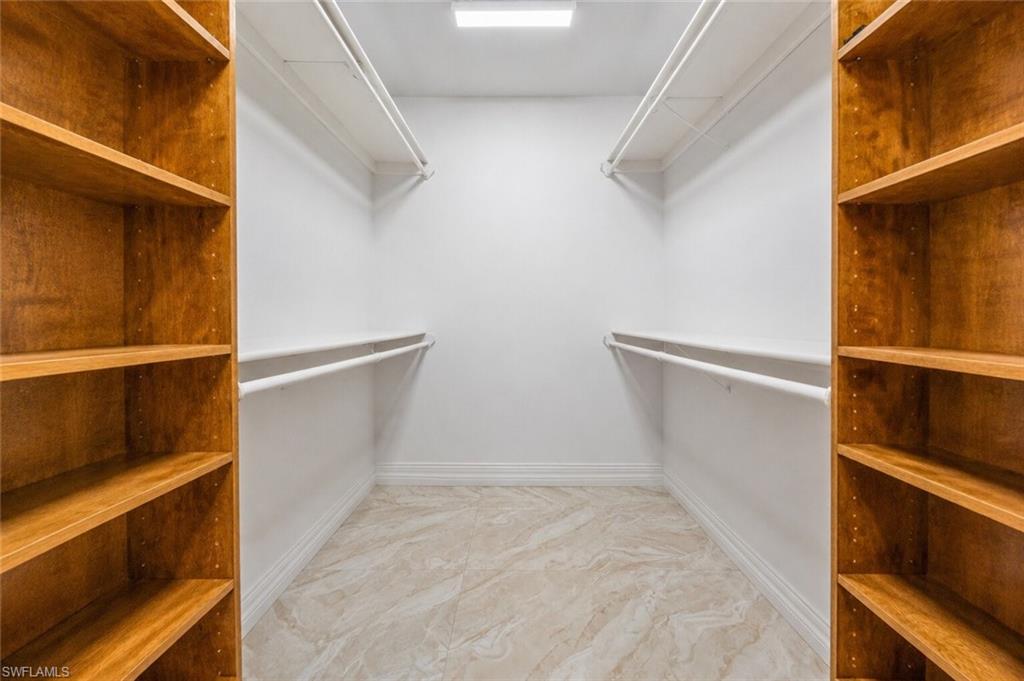Spacious walk-in closet featuring light wood-finish shelving units, white hanging rods, white baseboards, and light-toned tile flooring