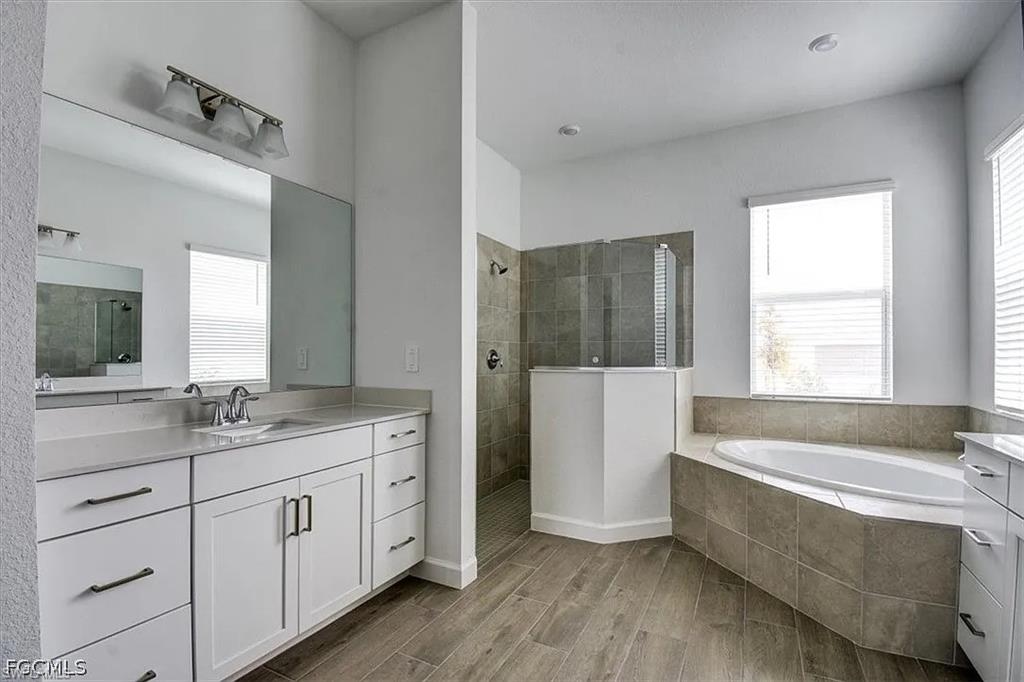 Bathroom featuring a large soaking tub, separate tiled shower with glass enclosure, wood-finish flooring, dual white vanities with integrated sinks, and ample natural light
