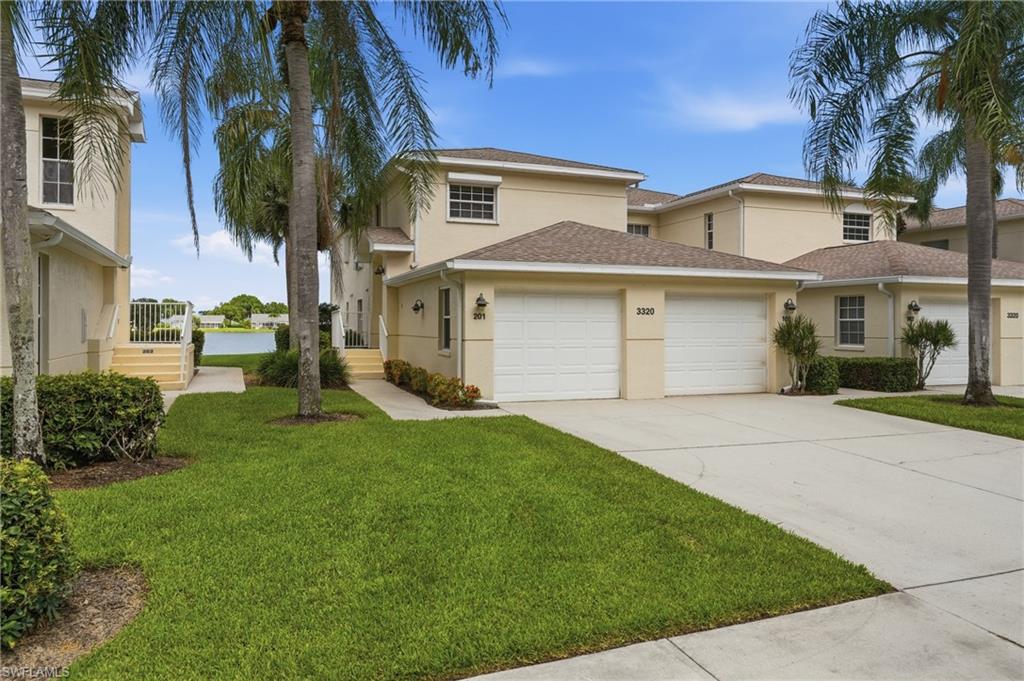 Contemporary stucco exterior featuring a two-car garage, mature palm trees, a concrete driveway, a manicured lawn, and a distant water body