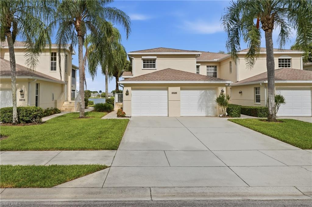 Cream stucco exterior with brown shingle roof, featuring two white panel garage doors, established palm trees, and manicured landscaping