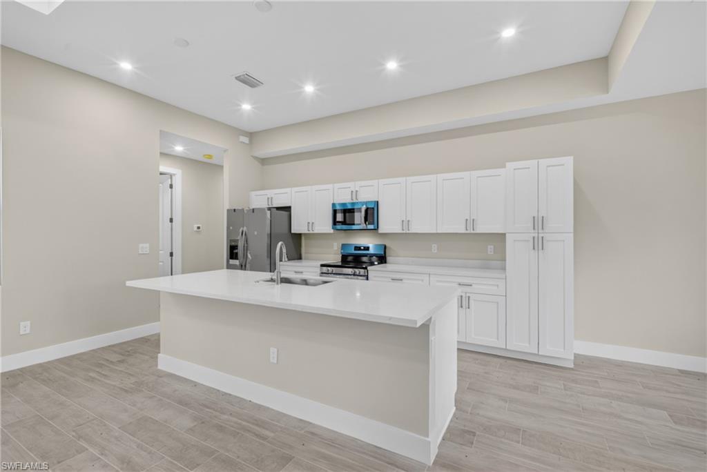 Bright kitchen featuring a central island with a sink, white cabinetry, integrated stainless steel appliances, and wood-finish flooring