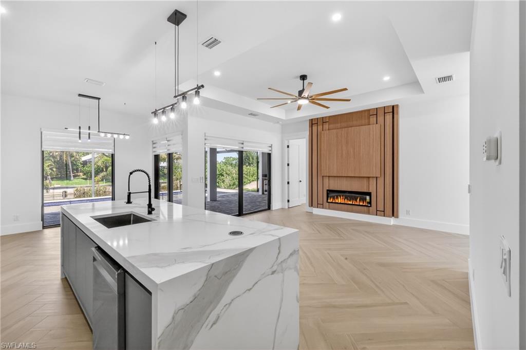 Open-concept kitchen and living area featuring a large kitchen island with a waterfall countertop, herringbone wood-finish flooring, a contemporary fireplace with a wood-paneled accent wall, and recessed lighting
