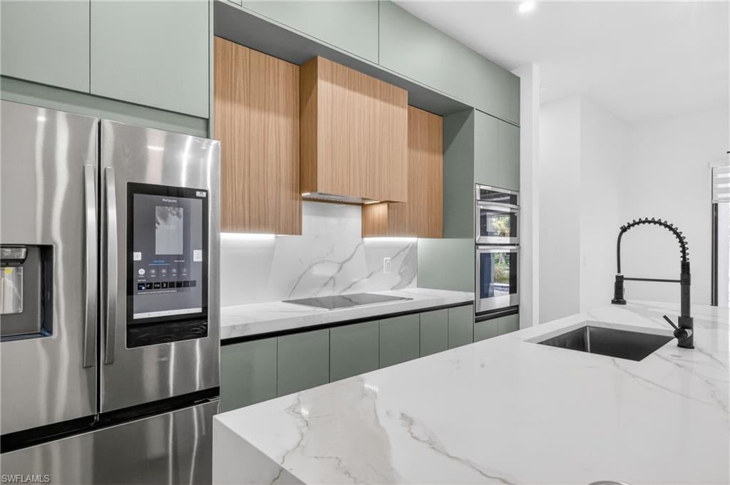 Kitchen featuring a built-in stainless steel refrigerator, double wall ovens, a black cooktop, a large island with an undermount sink, and wood-finish cabinetry accented with light green panels