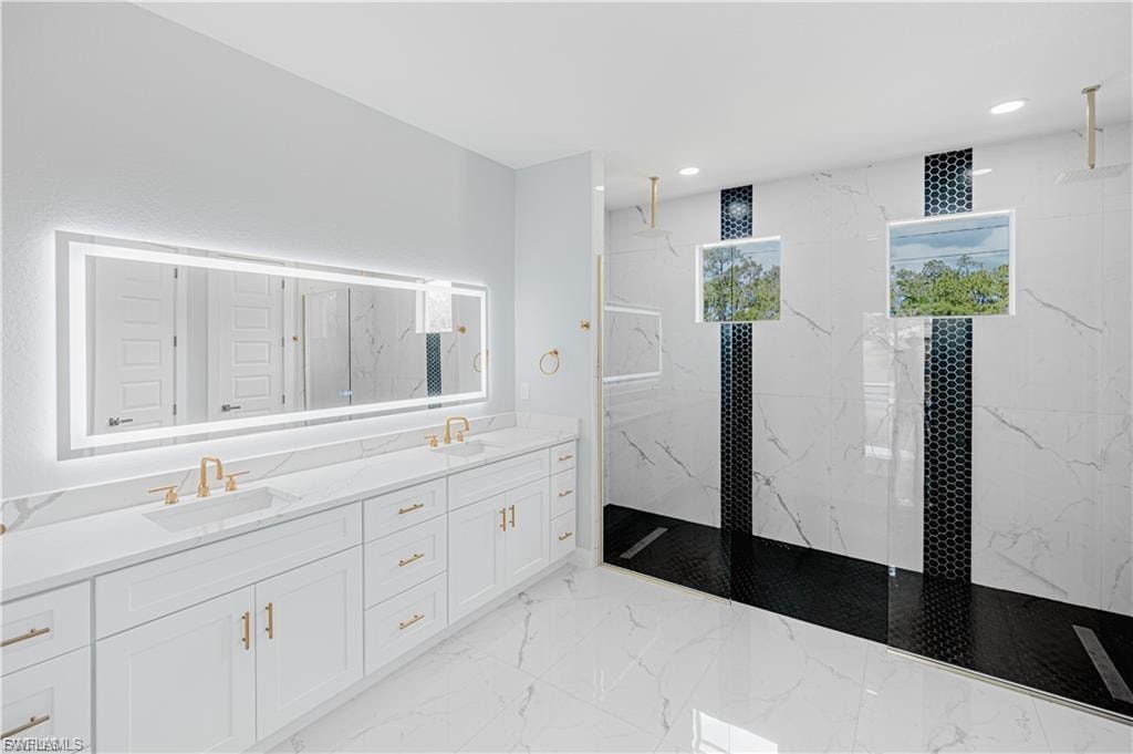 Spacious bathroom featuring a dual vanity with white cabinetry and gold-tone hardware, a large illuminated mirror, a walk-in shower with black hexagonal tile accents, and white veined tile flooring