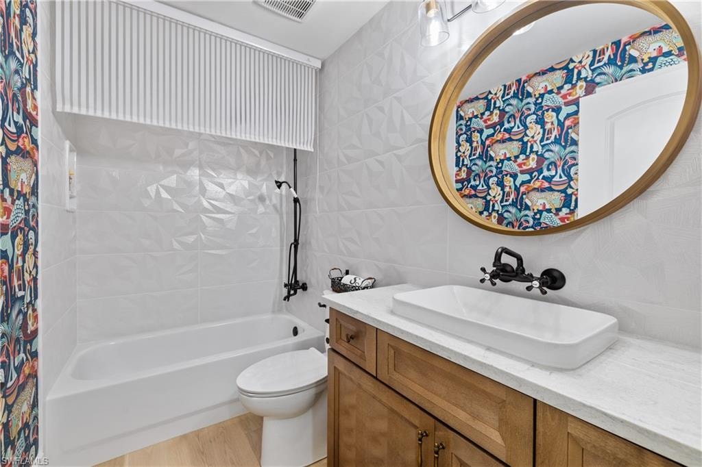 Bathroom featuring a built-in tub, white geometric-textured wall tiling, a wood-finish vanity with a vessel sink, a gold-framed circular mirror, and a wall-mounted matte black faucet