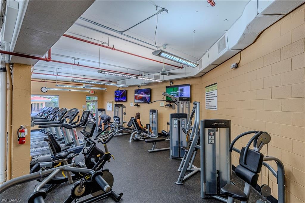 Fitness center featuring a selection of cardio equipment, strength training machines, multiple wall-mounted televisions, exposed ceiling ductwork, and fluorescent lighting