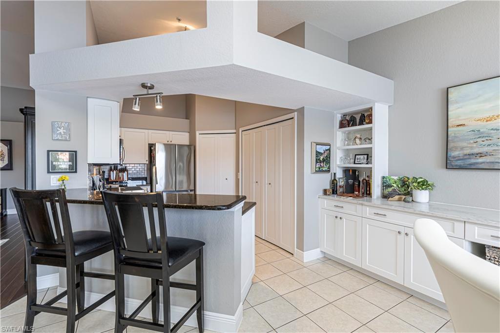 Open-concept living area featuring a kitchen island with a dark countertop, white shaker-style cabinetry, a built-in beverage bar with open shelving, and light-toned tile flooring
