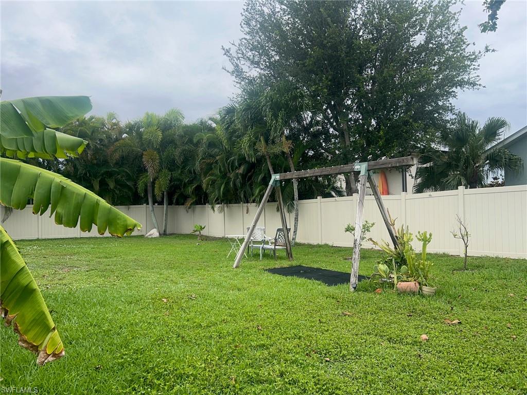Expansive backyard featuring a lush lawn, privacy fencing, mature trees, a wooden swing set, and a variety of potted plants