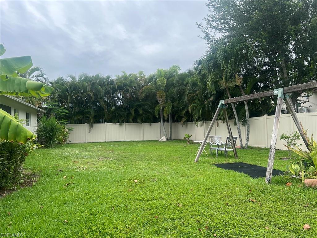 Expansive green lawn area featuring a tall privacy fence, mature tropical foliage, and a wooden playset