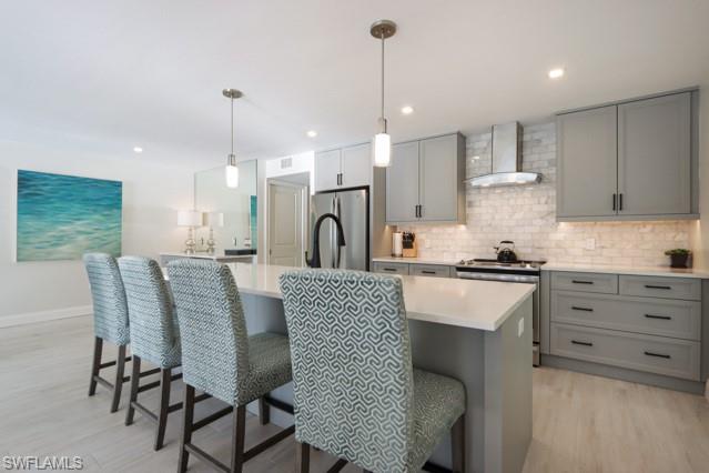 Kitchen with gray cabinets, appliances with stainless steel finishes, light wood-style flooring, tasteful backsplash, and wall chimney range hood