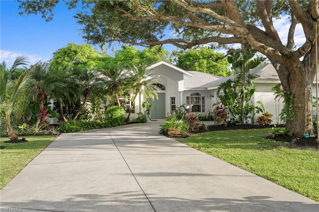 Front exterior featuring a concrete driveway, mature trees, manicured lawn, and lush landscaping