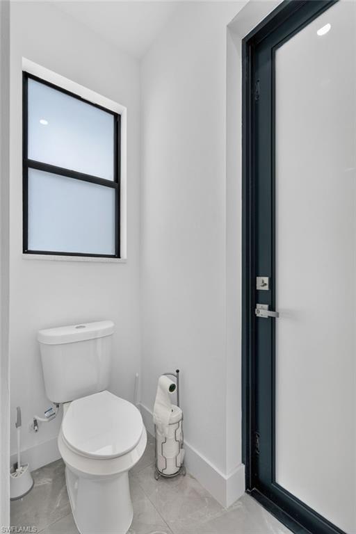 Compact bathroom featuring a white toilet, frosted-pane window with black framing, sleek black door frame, white baseboards, and grey tile flooring