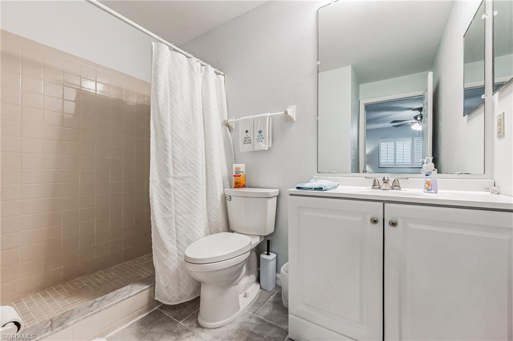 Bathroom featuring a single vanity with storage, a wall-mounted mirror, a commode, and a shower with tiled walls and a marble-finish threshold