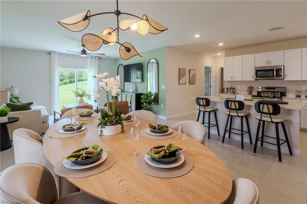 Open-concept living space featuring a dining area with a distinctive chandelier, a contemporary kitchen with white cabinetry, a kitchen island with seating, stainless steel appliances, and a living area with sliding glass doors opening to a green exterior