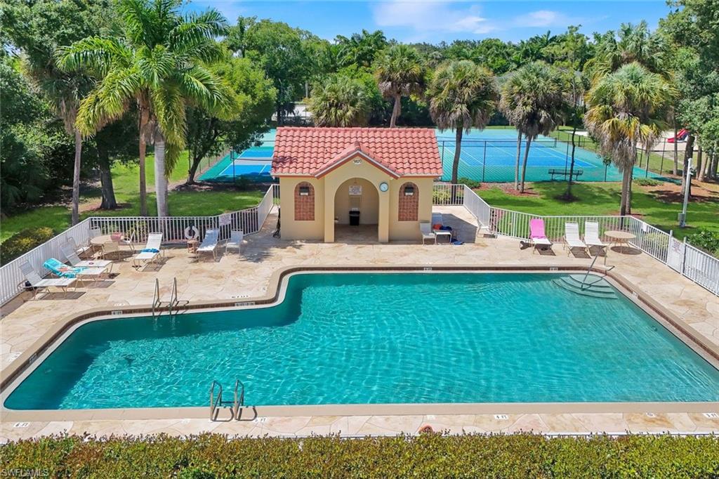 Shaped swimming pool with a surrounding paver deck, a pool house featuring a red tile roof, and a white perimeter fence