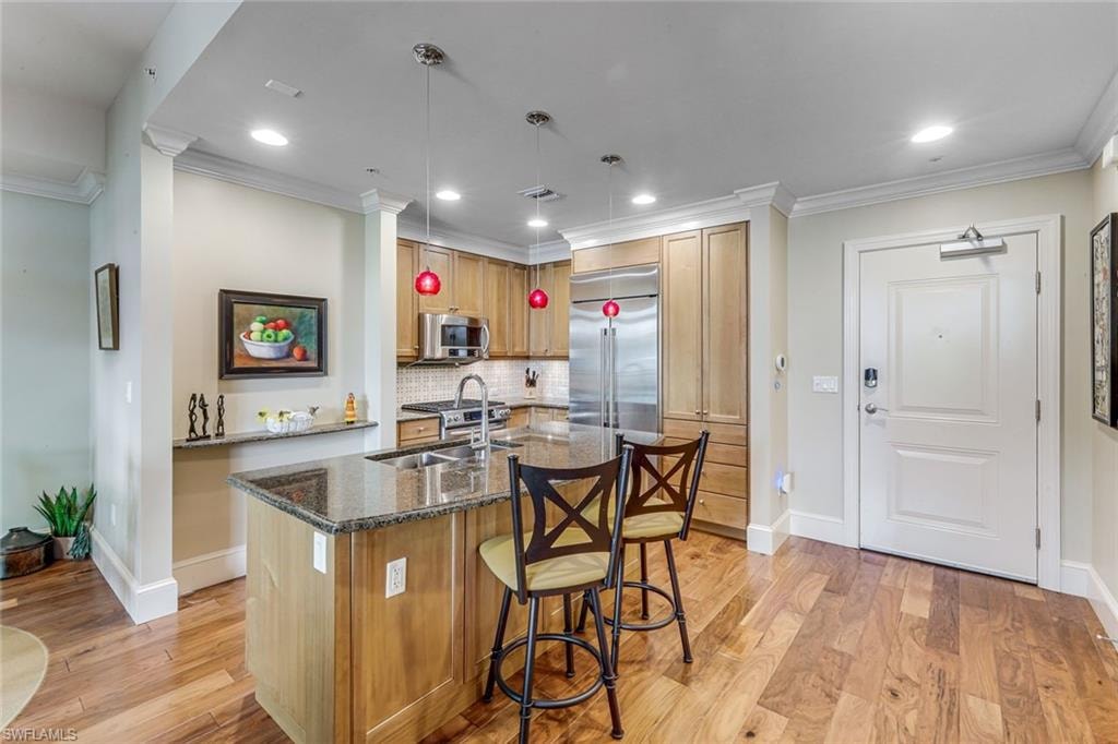 Kitchen featuring a central island with a granite countertop and integrated sink, stainless steel appliances, light wood cabinetry, and wood-finish flooring