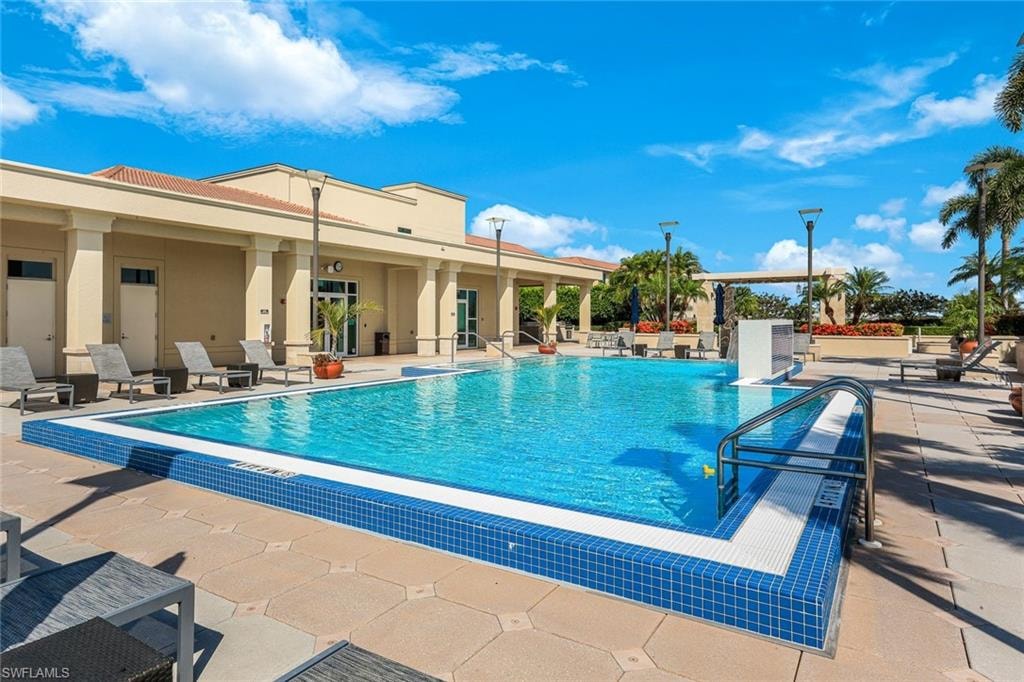 Resort-style pool area featuring a blue mosaic tile border, concrete paver decking, a light-toned stucco building with columns, and surrounding tropical landscaping including palm trees