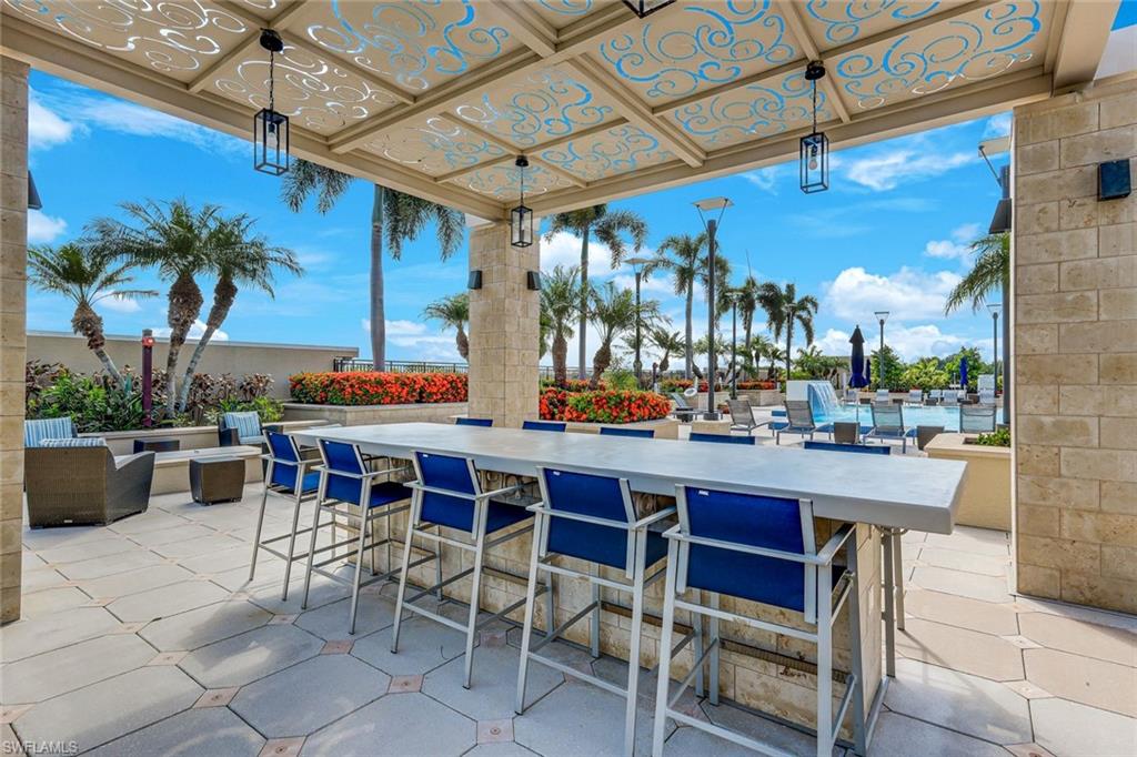 Covered outdoor patio featuring a patterned overhead trellis with pendant lighting, a long bar area with seating, and stone tile flooring