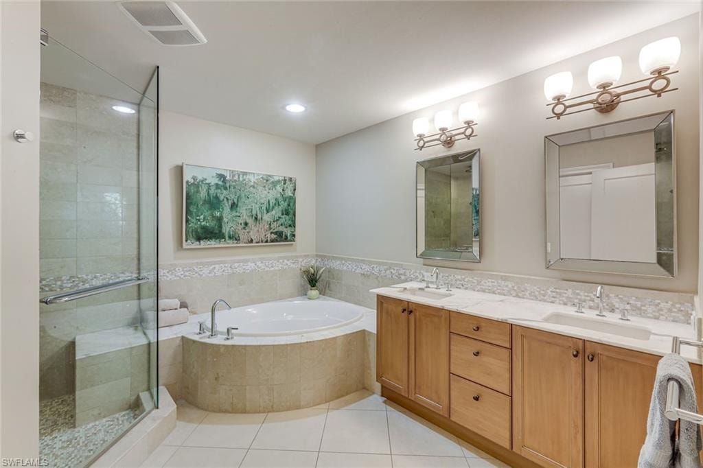 Tiled bathroom featuring a glass-enclosed shower with bench seating, a separate soaking tub, and a dual vanity with light-colored wood cabinetry