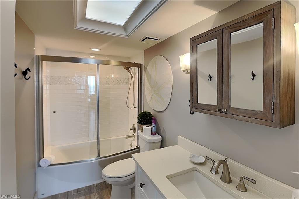 Bathroom featuring a white subway tile tub/shower combination with sliding glass doors, a vanity with a white countertop and undermount sink, wood-finish flooring, a wood-framed medicine cabinet with mirrored doors, and a skylight