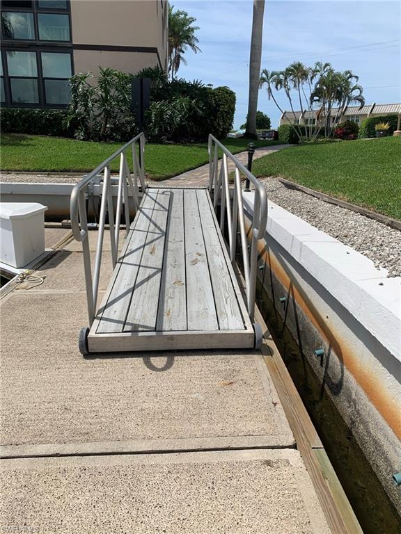 Waterfront access featuring a wooden dock with metal handrails, a concrete walkway, mature palm trees, and manicured green landscaping