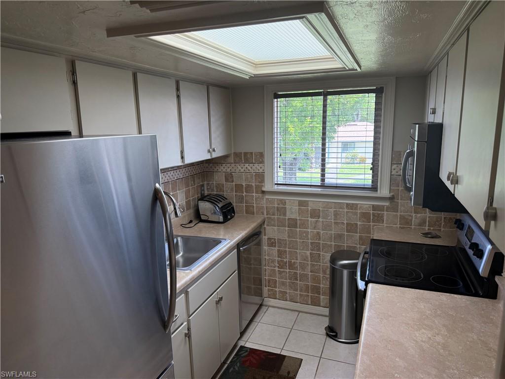 Galley kitchen featuring a skylight, window with blinds, stainless steel appliances, white cabinetry, and a tile backsplash