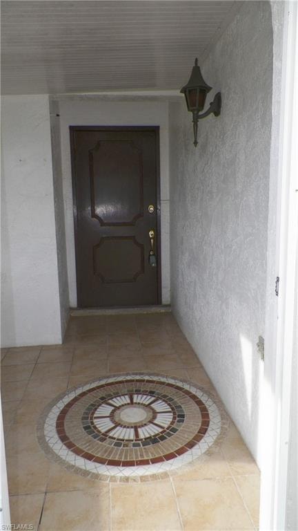 Entryway featuring a decorative mosaic tile floor medallion, a dark wood-finish entry door, textured exterior wall finish, and an outdoor wall sconce
