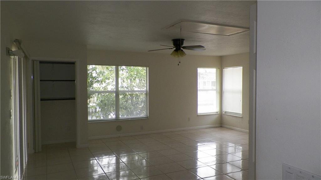 Spacious room featuring light-toned square tile flooring, multiple windows providing ample natural light, a ceiling fan with integrated lighting, and a built-in closet with shelving