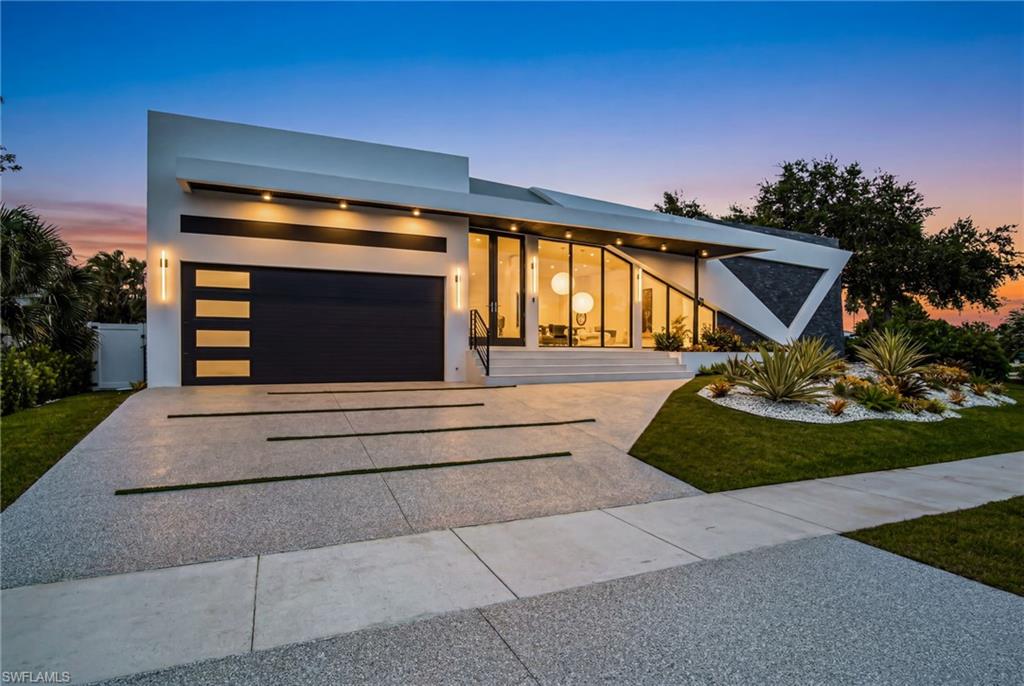 Modern architectural exterior featuring a distinct angular design, expansive glass paneling, a two-car garage with frosted glass inserts, a decorative stone facade, and a meticulously landscaped front yard with white stone accents