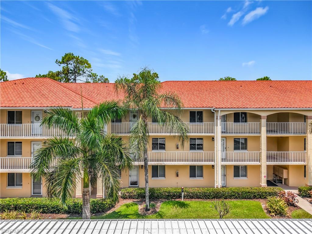Multi-story building featuring a terracotta tile roof, multiple balconies with white railings, a stucco exterior, and mature palm trees