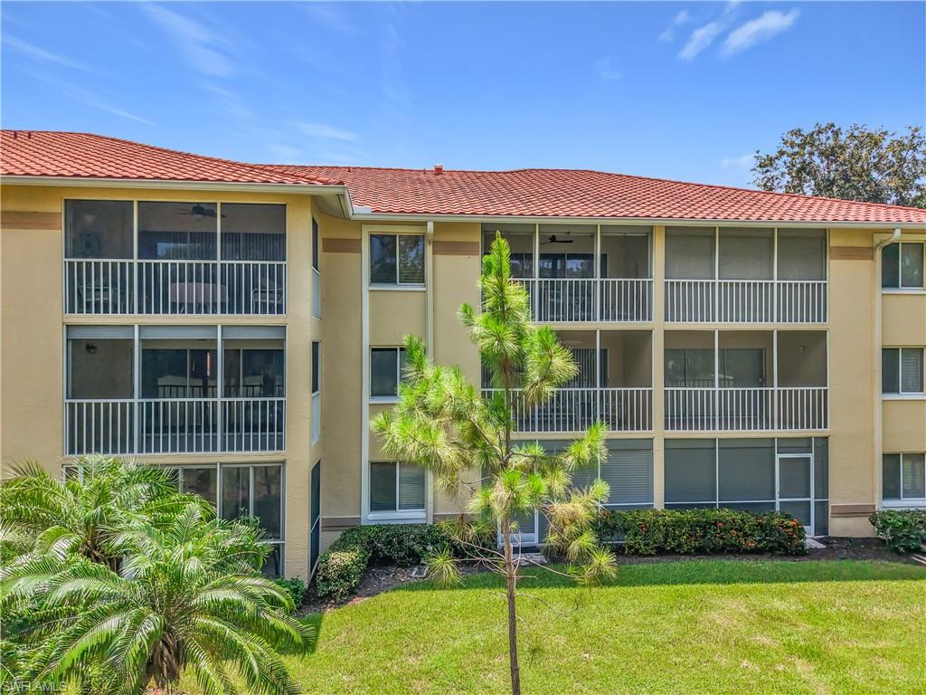 Multi-story building featuring screened balconies, a red tile roof, beige exterior siding, and surrounding green landscaping