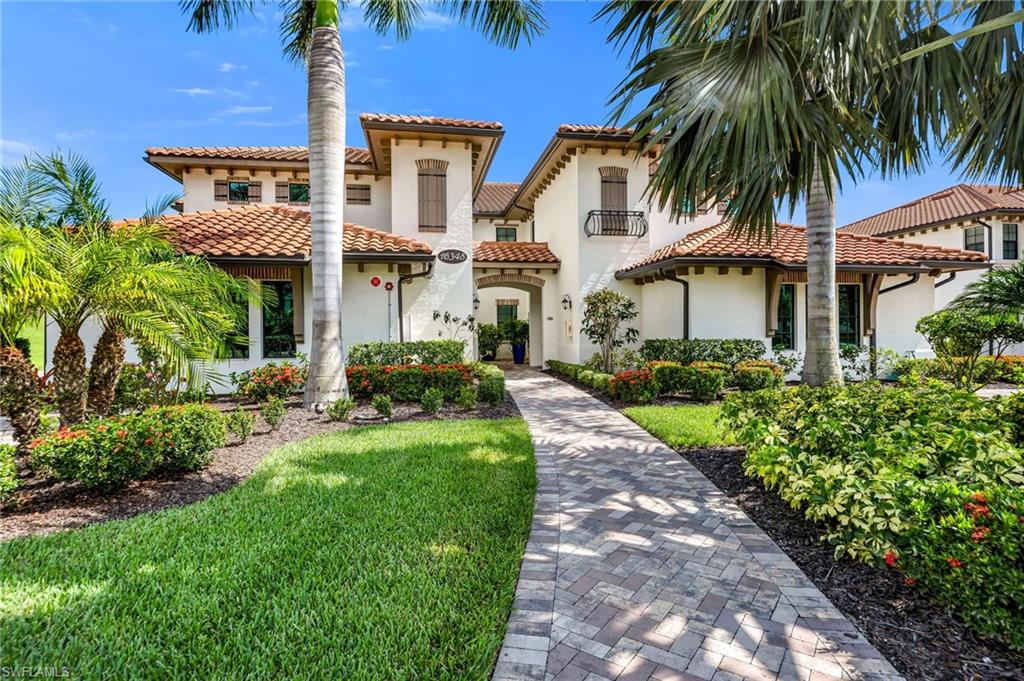 Mediterranean-inspired exterior featuring a paver walkway, mature palm trees, lush landscaping, and a terra cotta tile roof