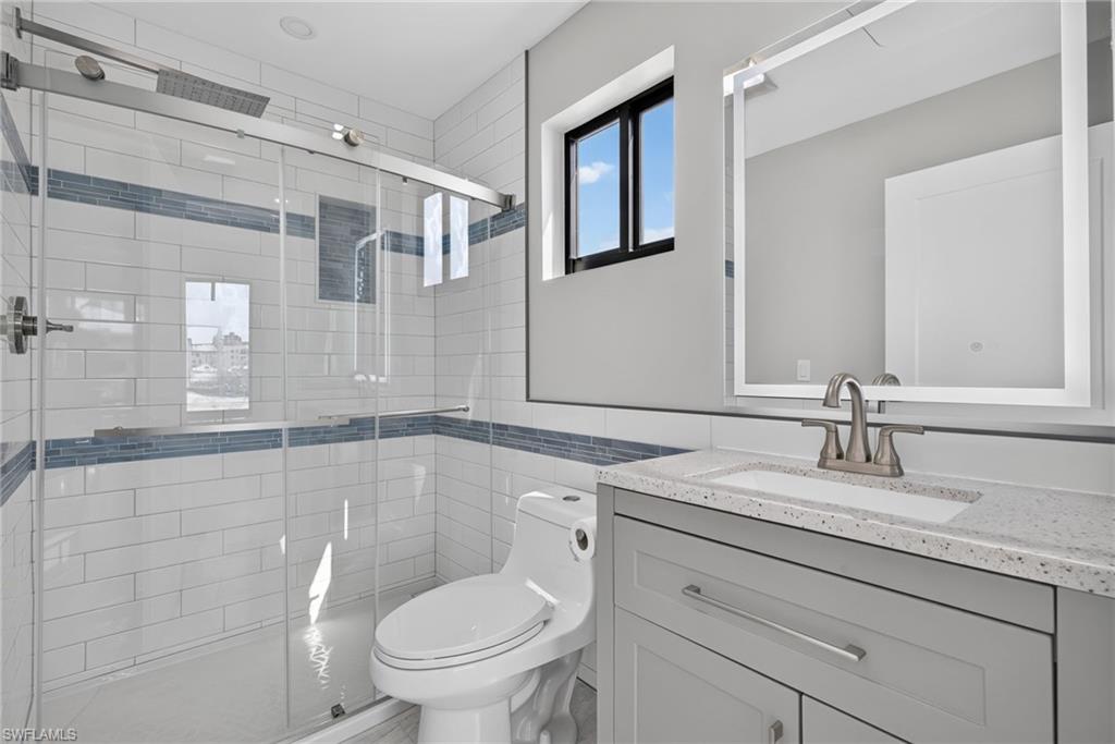 71 Mango Unit 1 Contemporary bathroom featuring a glass-enclosed shower with subway tile surround and integrated niche, a single-sink vanity with light gray cabinetry and a speckled countertop, a rectangular mirror, and a black-framed window