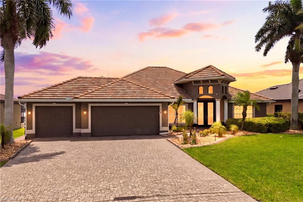Mediterranean-inspired facade featuring a tiled roof, paver driveway, two-car garage, manicured lawn, and tropical landscaping