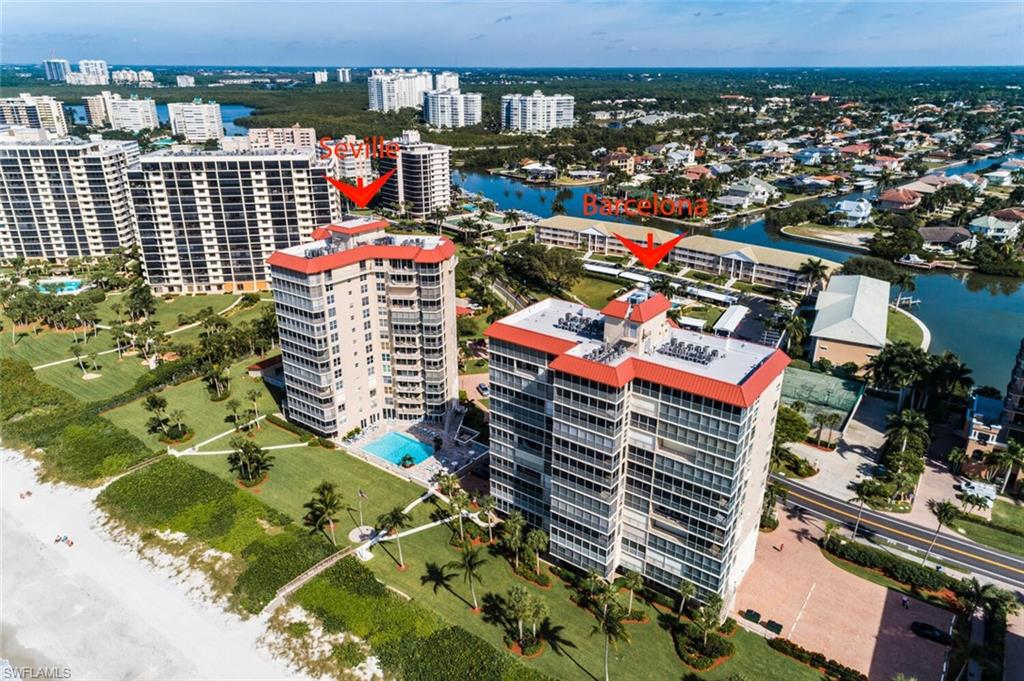 Aerial depiction of multiple high-rise buildings with red-tiled roofs along a coastline, featuring a beachfront, manicured green spaces, and a swimming pool