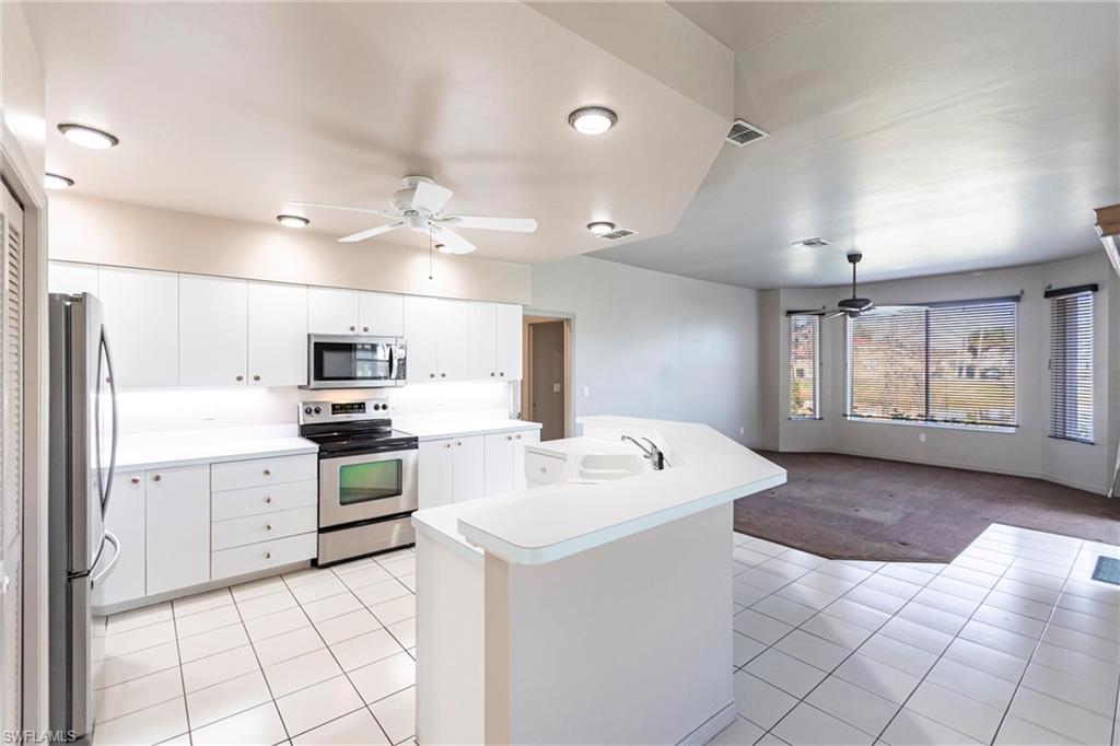 Kitchen with breakfast bar & dining area, white cabinetry, sink, light tile patterned floors, ceiling fan, and stainless steel appliances. View of carpeted entertainment area overlooking lake with bay window (manual shutters on all 3 windows), custom blinds, too!