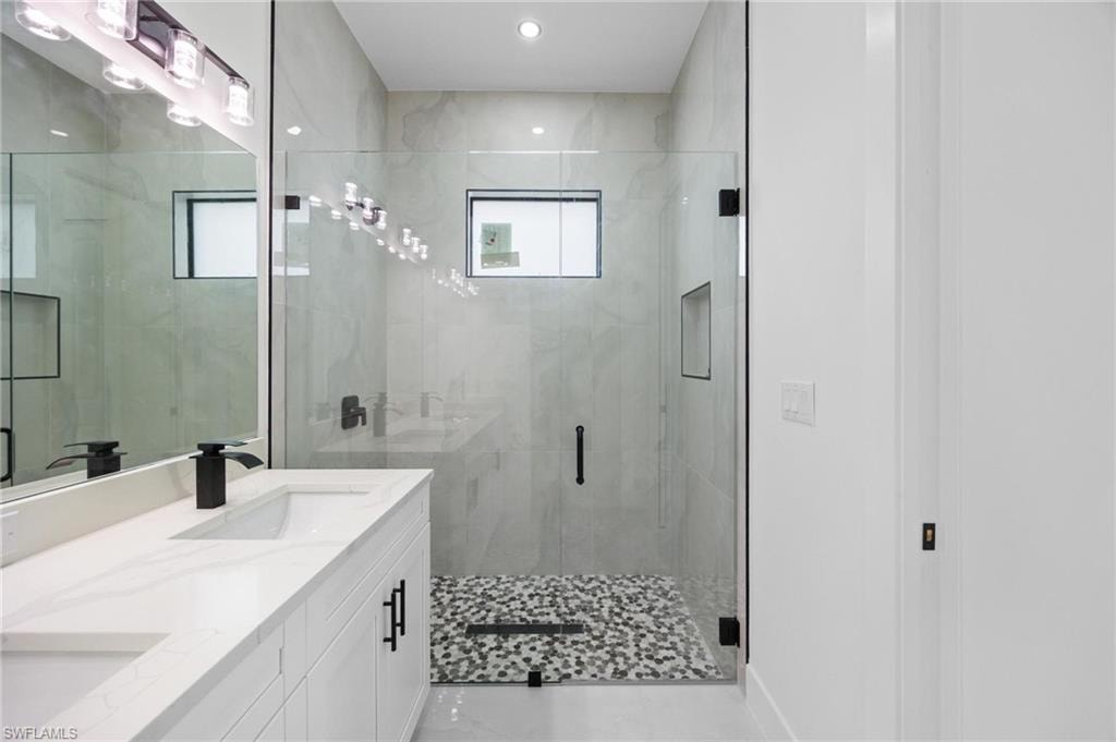 Spacious bathroom featuring a frameless glass shower enclosure, mosaic pebble floor, tiled shower walls, dual vanity with quartz countertop, and black hardware accents