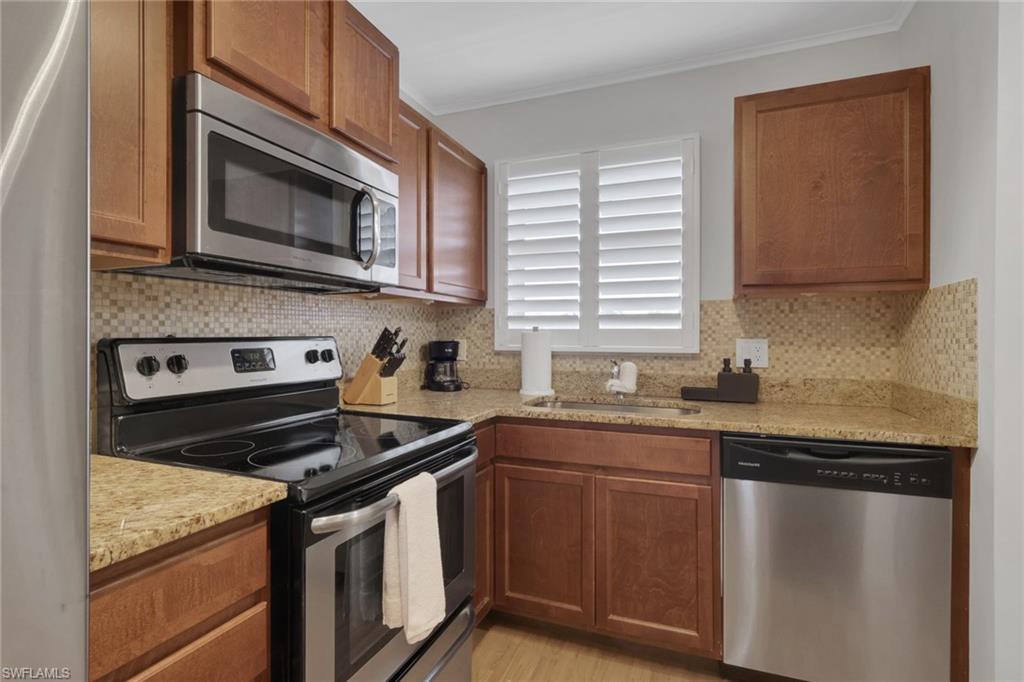 Kitchen featuring wood cabinetry, stainless steel appliances, granite-finish countertops, mosaic tile backsplash, and a window with white plantation shutters