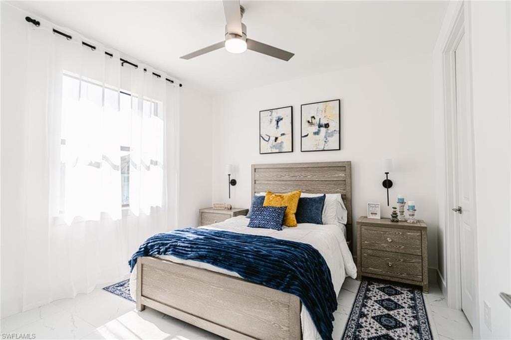 Bright bedroom featuring a large window with sheer white curtains, a ceiling fan with integrated lighting, and white flooring