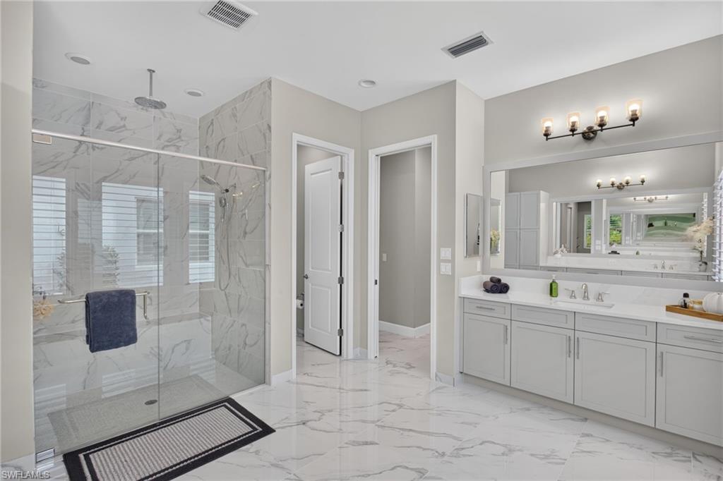 Spacious bathroom featuring marble-look flooring and shower surround, a glass-enclosed shower with a rainfall showerhead, and a double vanity with grey cabinetry and white countertops