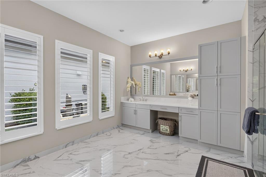 Spacious bathroom featuring large format marble-look tile flooring, light gray cabinetry, an expansive vanity with double sinks, a full-width mirror, and three windows with white plantation shutters