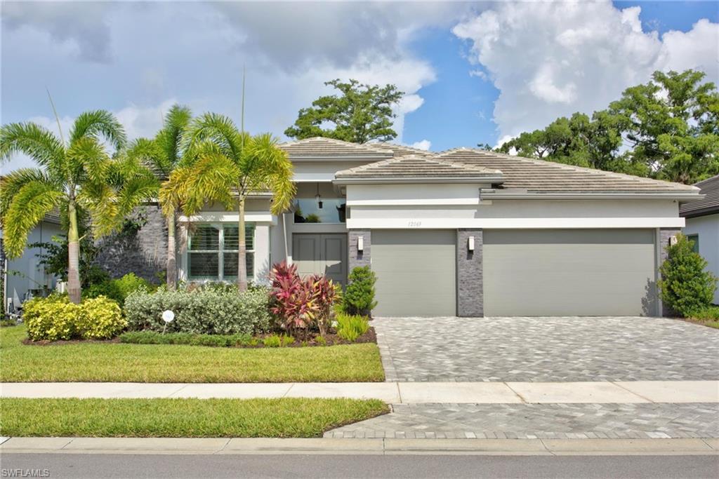 Contemporary stucco exterior with a tiled roof, paver driveway, two-car garage, and established landscaping featuring palm trees
