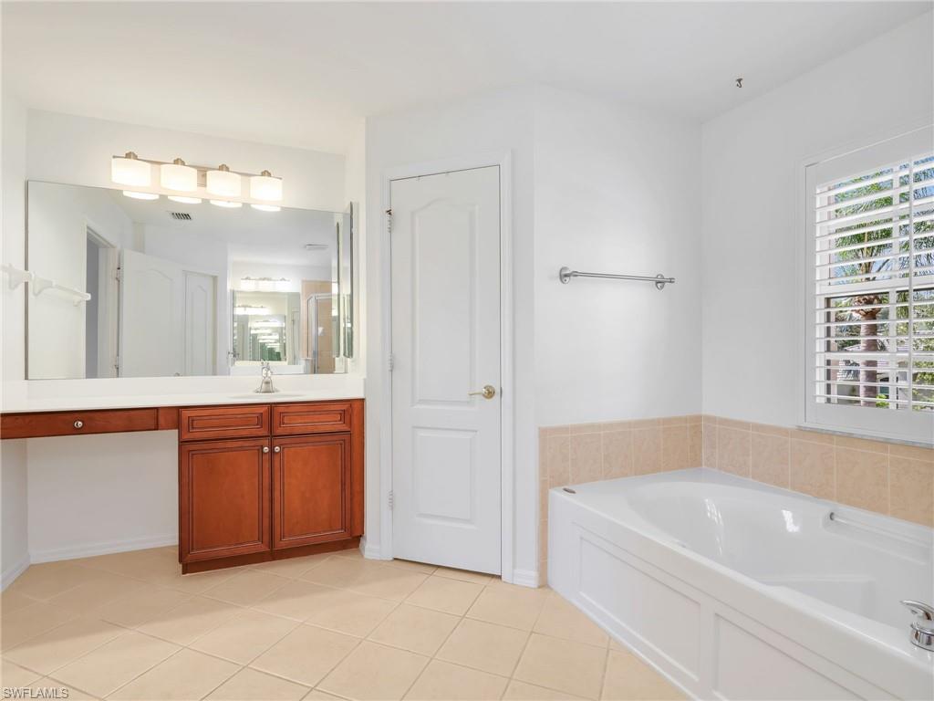 Bathroom featuring a built-in soaking tub with tiled surround, plantation shutters, and a vanity with a white countertop and wood-finish cabinetry